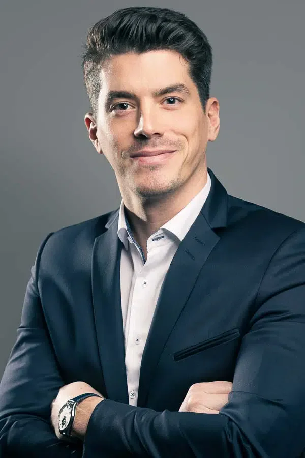 Portrait professionnel d'un homme d'affaires confiant, en costume foncé, souriant et croisant les bras, fond gris.